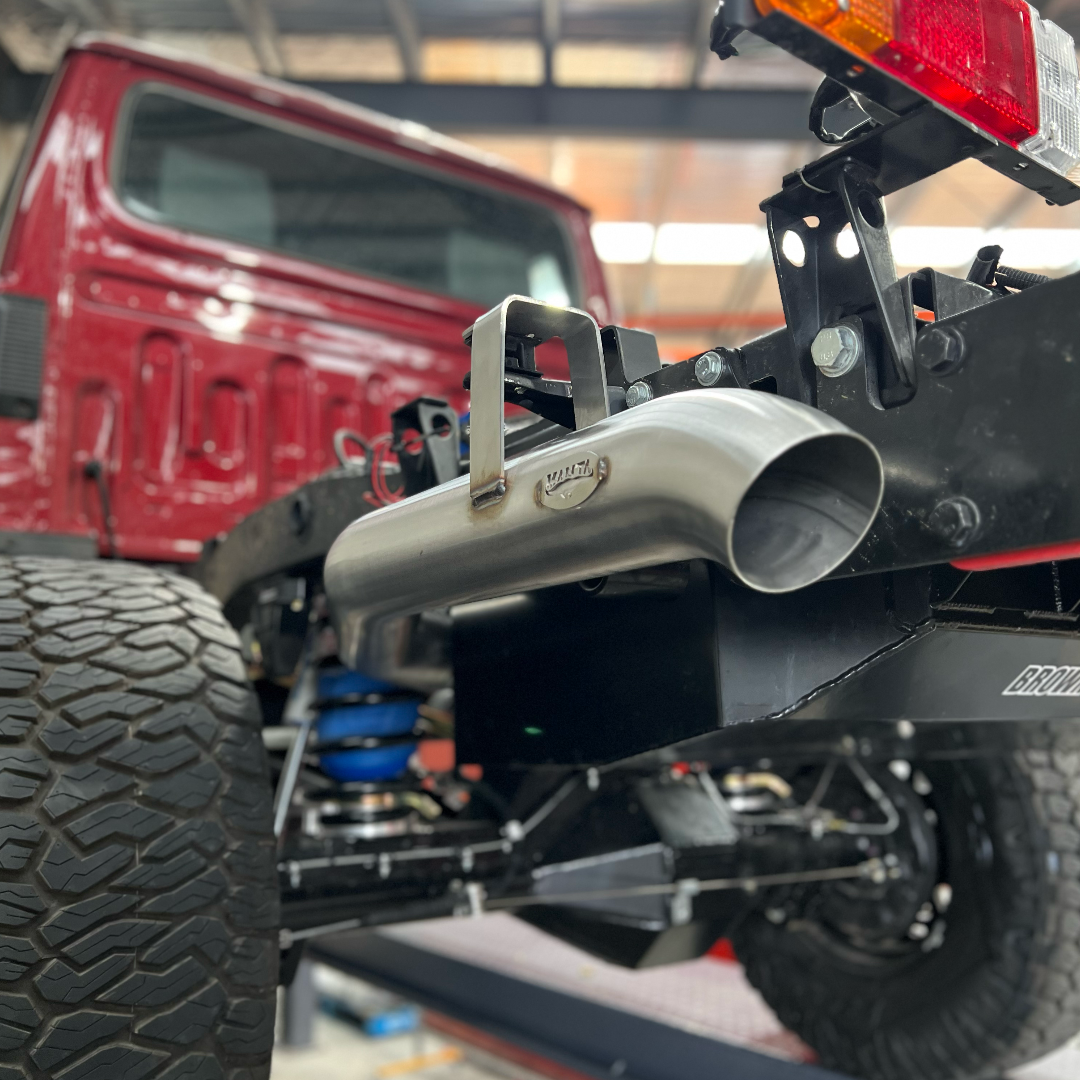Close-up of a red trucks unfinished rear, showing a large off-road tire, exposed suspension, and a polished exhaust pipe against a garage backdrop.
