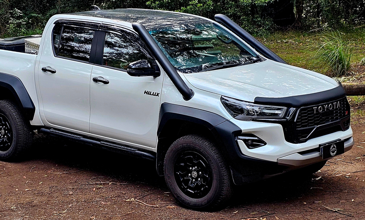 White Toyota Hilux with black accents parked on a dirt path surrounded by greenery.