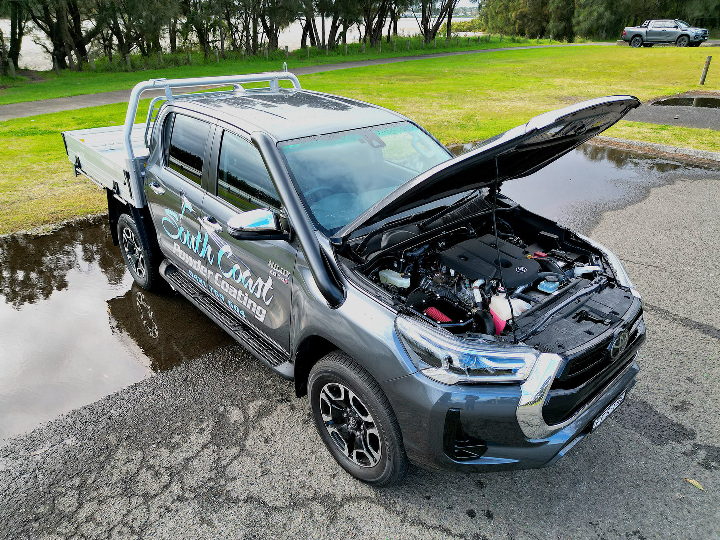 A gray pickup truck is parked on wet pavement with its hood open, revealing the engine.