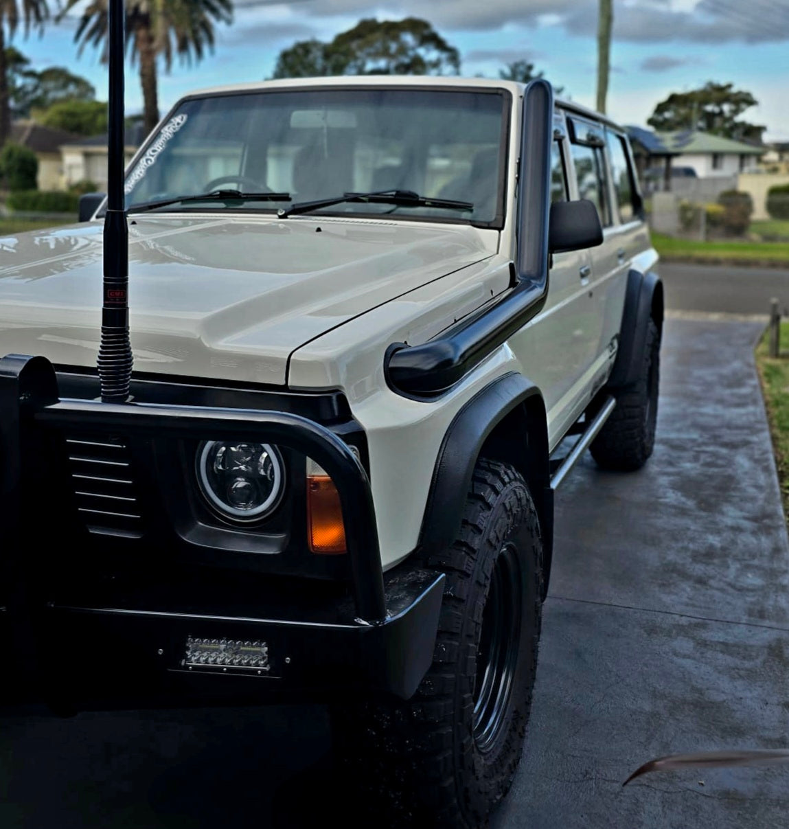 A white off-road vehicle with large tires and a snorkel attachment is parked on a driveway.