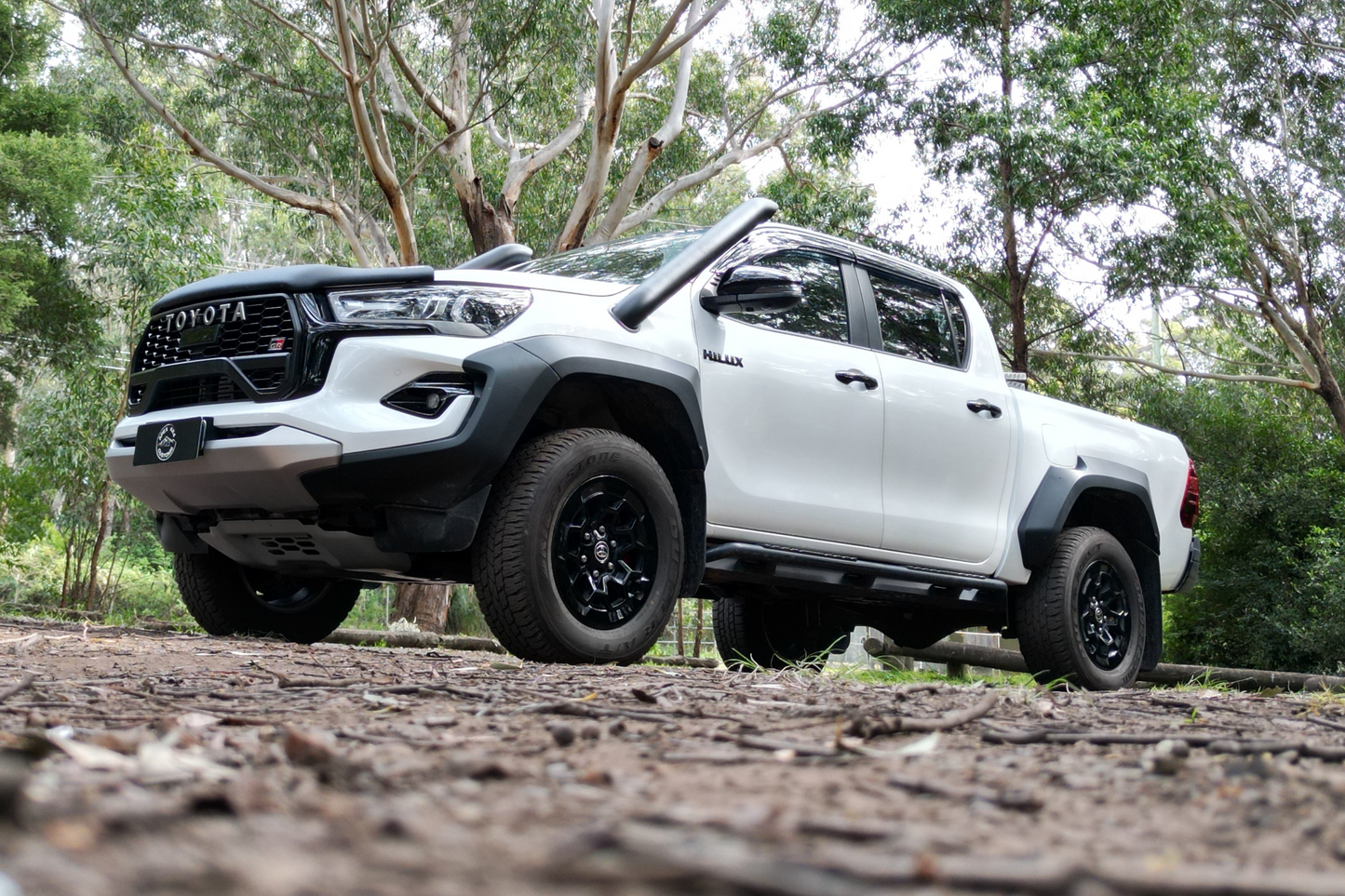 A white Toyota pickup truck with high ground clearance and aggressive styling is parked on a forest trail, surrounded by tall trees and greenery.